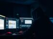 Eine Person mit Kapuze sitzt in einem dunklen Raum vor mehreren Monitoren, auf denen das Microsoft-Logo und eine Phishing-Nachricht zu sehen sind.