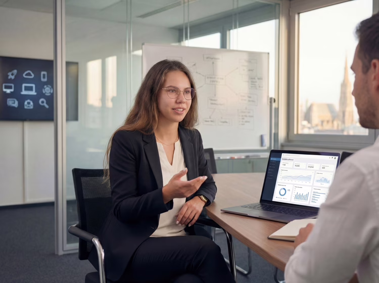 IT consultant Daniela Söldner advising a client in a modern Vienna office, with analytics dashboard on laptop and IT infrastructure diagram on whiteboard, Vienna skyline visible through window